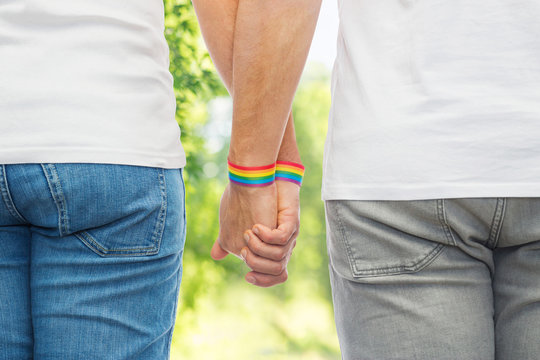 lgbt, same-sex relationships and homosexual concept - close up of male couple wearing gay pride rainbow awareness wristbands holding hands over green natural background