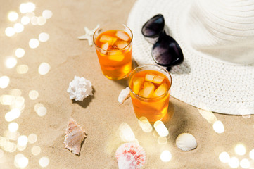 Obraz na płótnie Canvas vacation, travel and summer concept - two glasses of aperitif cocktails with ice cubes, sun hat, sunglasses and seashells on beach sand