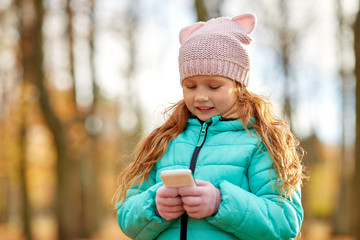 childhood, season and people concept - happy girl with smartphone at autumn park