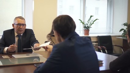 Negotiation briefing with multiethnic partners in office conference room. Lawyer giving contract draft to mature businessman and african-american businesswoman. Slow motion.