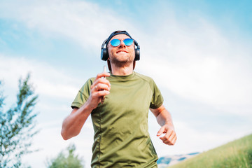 Bearded man in baseball cap, wireless headphones and blue sunglasses cheerful smiling running evening run and holding in hand modern smartphone device.