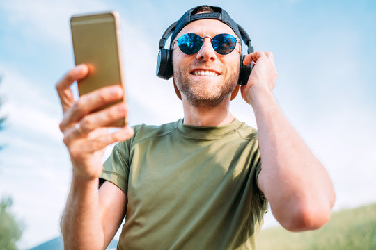 Cool man in baseball cap, wireless headphones and blue sunglasses browsing in his mp3 player playlists smartphone device and enjoying favorite music playing.