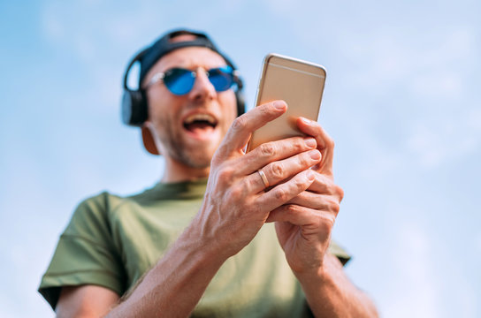 Cool man in baseball cap, wireless headphones and blue sunglasses browsing in his mp3 player playlists smartphone device and cheerful singing a favorite song.