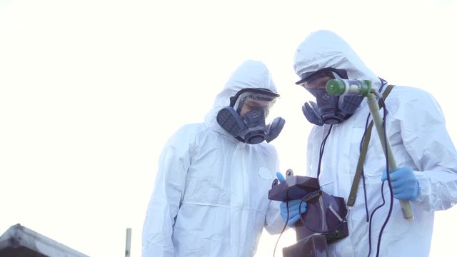 two scientists in protective suits and masks and make measurements of radiation against the background of the ruins at sunset