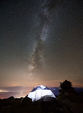 Tourist camping at summer night on the top of rocky mountain. Glowing tent under incredibly beautiful night sky full of stars and Milky way. On background starry sky, mountains and luminous town