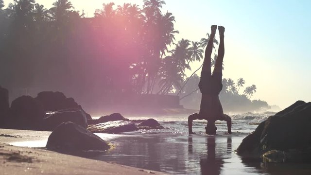muscular athlete stands on hands and head in shallow ocean water in sunny summer morning slow motion. Concept fitness and healthy lifestyle