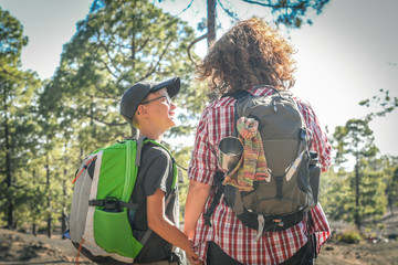 Fototapeta na wymiar Back view of mother and son trekking in a mountain forest. Mum and child with backpack hiking into the park. Smiling boy looking at her mummy. Travel adventure health and togetherness concept.