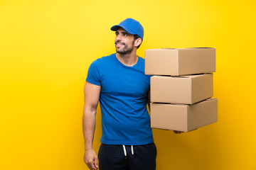 Young delivery man over isolated yellow wall looking up while smiling