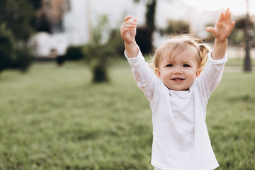 little baby girl with blond hair and two tails, wearing a white T-shirt standing on green grass in nature, smiling
