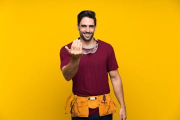 Handsome young craftsman over isolated yellow background inviting to come with hand. Happy that you...