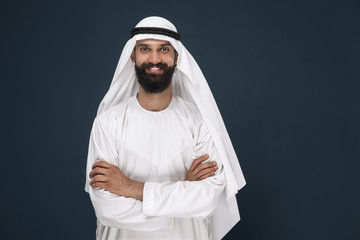 Half-length portrait of arabian saudi businessman on dark blue studio background. Young male model standing and smiling. Concept of business, finance, facial expression, human emotions.