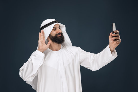 Half-length Portrait Of Arabian Saudi Man On Dark Blue Studio Background. Young Male Model Using Smartphone, Making Selfie. Concept Of Business, Finance, Facial Expression, Human Emotions