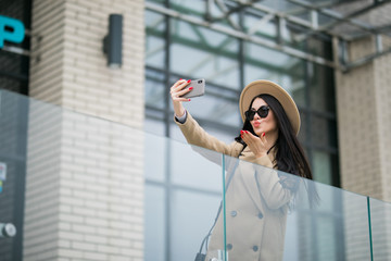 Portrait of pretty young woman making selfie by the phone. Smiling, walking on the city street. Going shopping. Wearing stylish white coat.