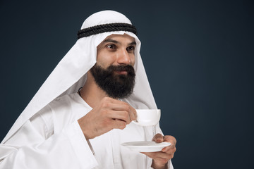 Half-length portrait of arabian saudi businessman on dark blue studio background. Young male model standing and drinking coffee or tea. Concept of business, finance, facial expression, human emotions.