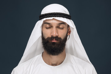 Half-length portrait of arabian saudi businessman on dark blue studio background. Young male model with closed eyes thinking. Concept of business, finance, facial expression, human emotions.