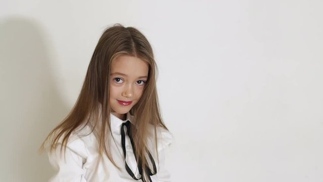 A cute teen girl in a white blouse with a black tie and long hair posing in the Studio on a white background, she straightens her hair with her hands. Slow motion.
