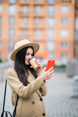 Cheerful young woman wearing coat using her phone in the sunny city street and drinking take away...