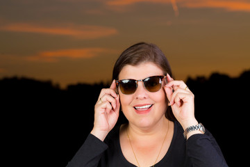 Woman Portrait with a big smile holding sunglasses in hand during twilight that is behind her