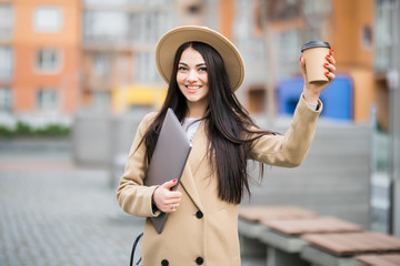 Pretty young woman holding laptop and a cup of coffee and files walking on the street