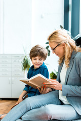 woman in glasses sitting on floor and reading book with cute kid