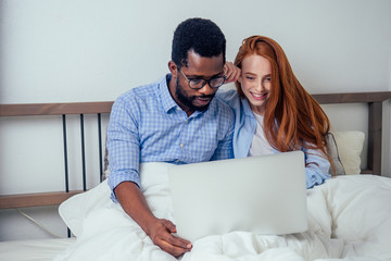 ginger redhaired european female and handsome afro african male together hugging lying in bedroom at home cozy apartment and watching news on laptop.lifestyle interracial family