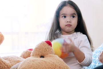 Cheerful cute little girl with big brown teddy bear sitting on bed while playing colorful ball in his hand at home. Lifestyle child relaxation concept