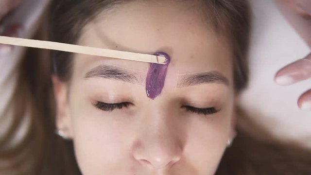 Young, caucasian girl is lying on couch during eyebrowes treatment at studio beauty, beautician depilating and shaping brows at beauty salon, removing the wax between the brows using a stick. Footage