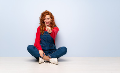 Redhead woman with overalls sitting on the floor showing and lifting a finger