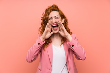 Redhead woman in suit over isolated pink wall shouting with mouth wide open