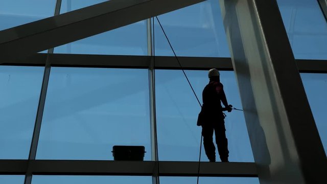 Worker man is washing windows with scraper on a highrise modern building hanging the rope, close-up