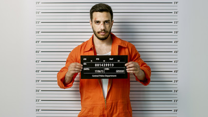 In a Police Station Arrested Man Getting Front-View Mug Shot. He's Wearing Prisoner Orange Jumpsuit and Holds Placard. Height Chart in the Background. - obrazy, fototapety, plakaty