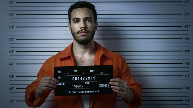 In a Police Station Arrested Man Getting Front-View Mug Shot. He's in a Prisoner Orange Jumpsuit and Holds Placard. Height Chart in the Background.