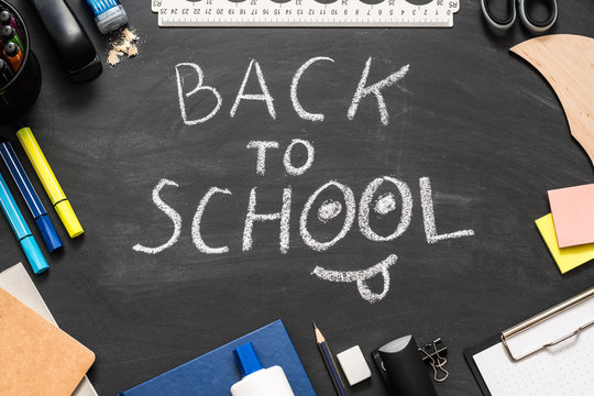 Education concept. Back to school white chalk lettering, smiley face over black chalkboard. Flat lay of stationery.