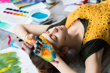 Art therapy. Tired young lady lying down on floor with her eyes closed, hands dirty with colorful paint.