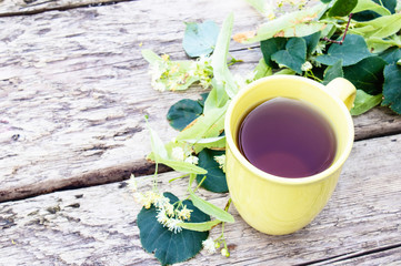 Linden tea in a yellow cup stands near linden flowers against the background of old boards.