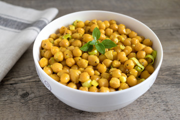 Cooked chickpeas with leek, chia seeds, sesame seeds and basil on top in white bowl. Wooden surface with kitchen towel.