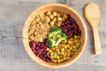 Vegan buddha bowl with stir fry tofu, brown rice, broccoli, red kidney beans, cooked chickpeas, seeds and vegetables. Flat lay on wooden surface with ladle.