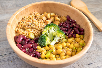 Vegan buddha bowl with stir fry tofu, brown rice, broccoli, red kidney beans, cooked chickpeas, seeds and vegetables. Wooden kitchen surface with ladle.