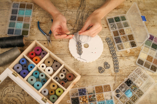 Top view of female hands making a macram&eacute; bracelet with kumihimo on a wooden table with tools, spools of thread, natural stones and colored beads