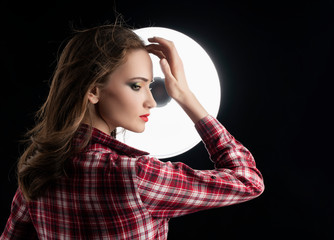 Beautiful girl model with red lips make-up, and hair flowing from the wind, wearing a casual plaid shirt near a round light on a black background. Advertising and fashion design.
