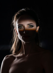 Beautiful girl model with red lips make up and naked shoulders in the shade, with a lit silhouette and a strip of light illuminating her eyes. Conceptual, art, fashionable design. Black background