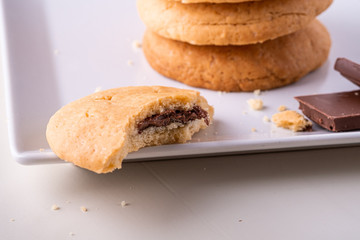 Homemade stack of shortbread chocolate cookies with one bitten cookie and two pieces of chocolate on white plate wooden table