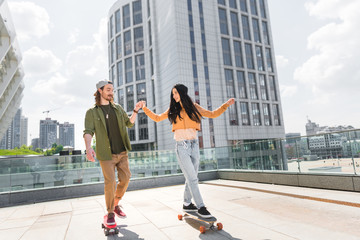 cheerful woman holding hands with man, riding on skateboard on roof