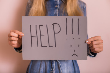 Woman holding paper with word help and sad face while standing in front of the wall.