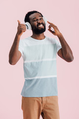 Fototapeta na wymiar cheerful african american man listening music with headphones and showing thumb up, isolated on pink