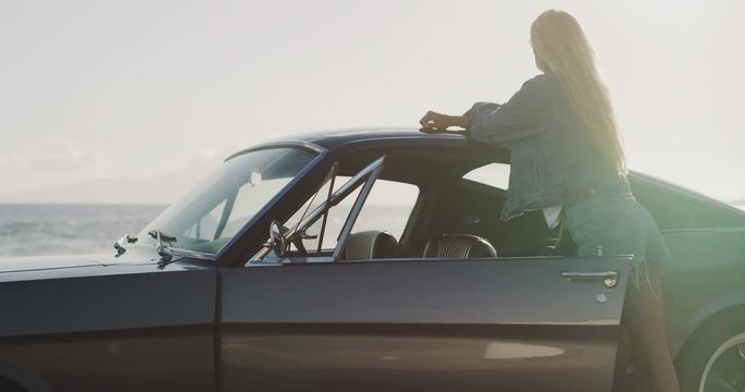 Attractive woman leaning on classic vintage sports car looking out at the ocean, woman going on a solo road trip