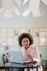 Beautiful mixed race woman in pink turtleneck sweater sitting in pastry shop and using laptop for on line shopping.