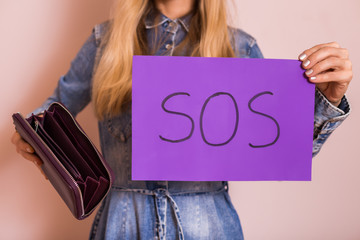 Woman holding empty wallet and SOS word written on paper while standing in front of wall.