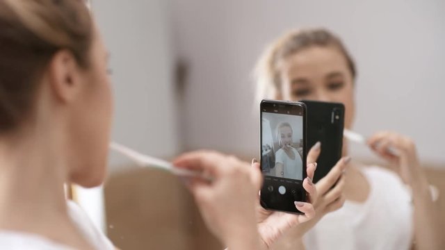 Over-shoulder shot of young Caucasian woman brushing her teeth in front of bathroom mirror and taking selfies on mobile phone, striking poses and pulling faces