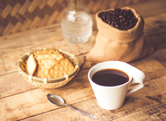 Black coffee In the cup white have roasted coffee beans are In a cloth bag sack butter cracker In the basket weave bamboo, and still have stainless steel spoon all placed on a wooden table.